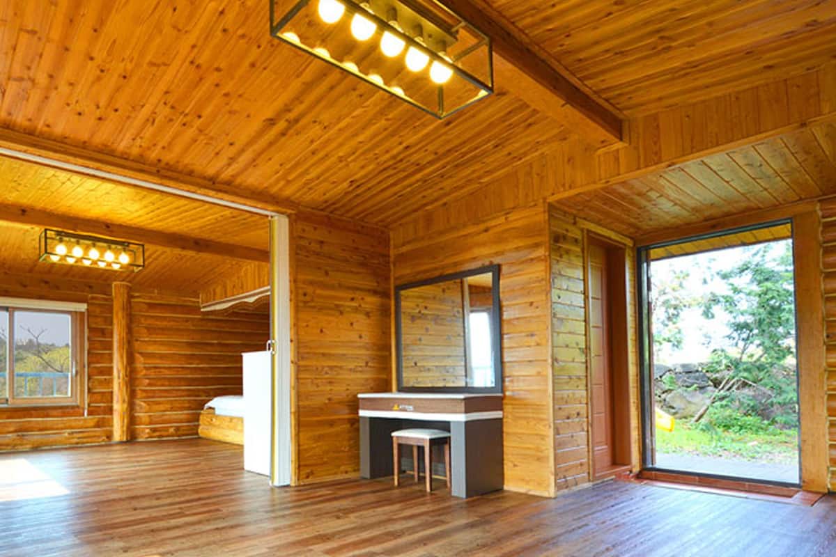 Interior de una cabaña de troncos, con piso de madera, un refrigerador blanco, un lavamanos con espejo y un ventanal con vistas al exterior.