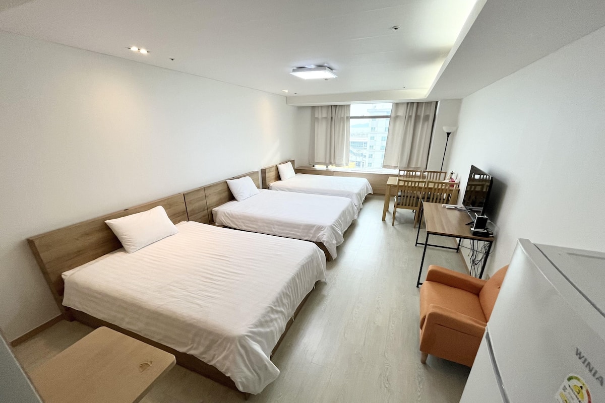 A hotel room with three single beds, a desk with a chair, a TV, and a window with curtains.