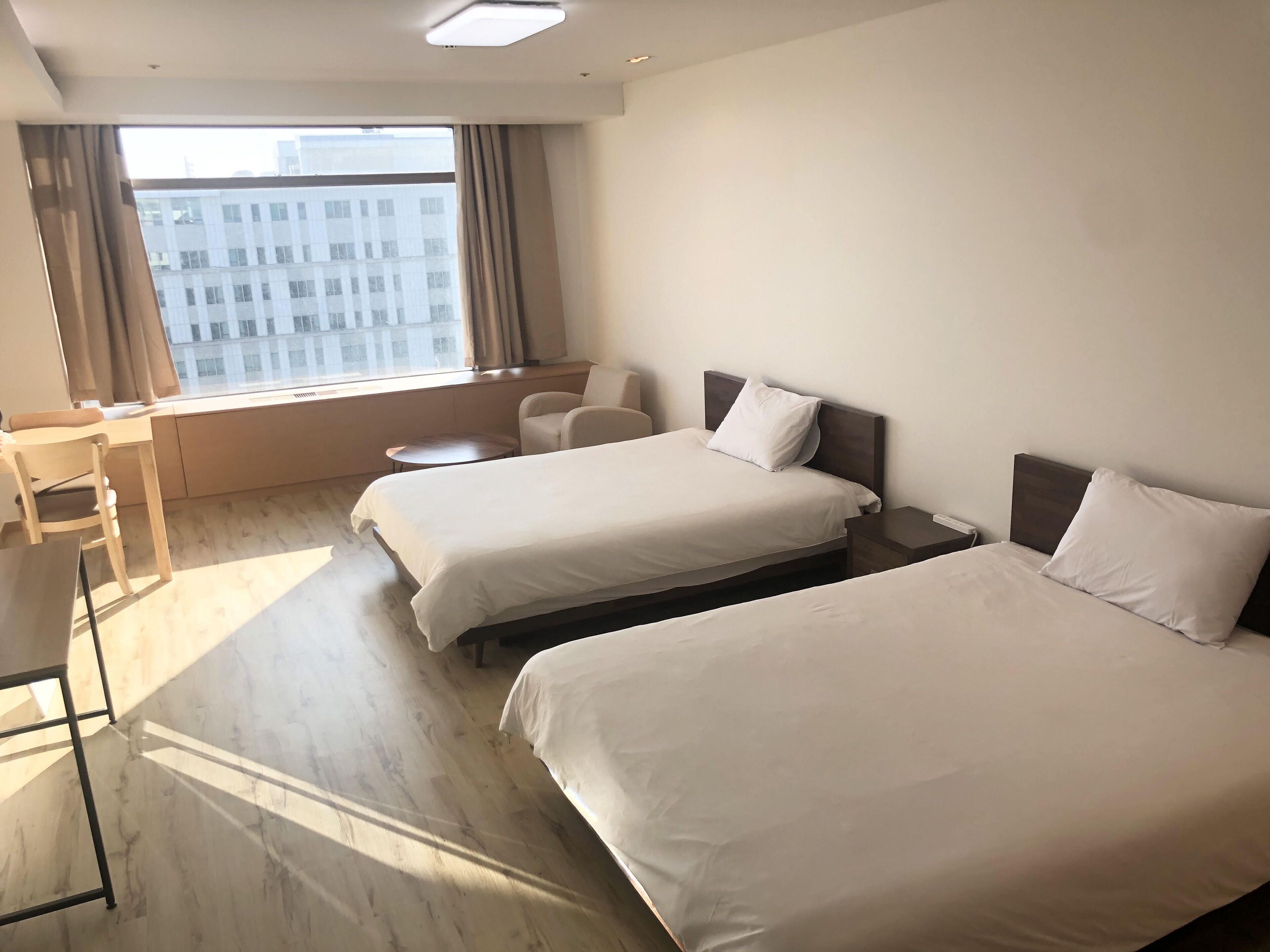 A hotel room with two beds, a desk, a chair, a small table, and a large window with a city view.