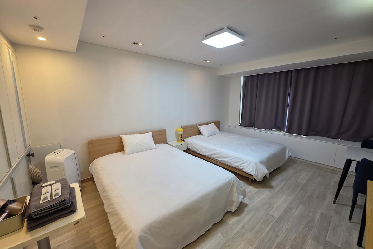 A hotel room with two beds, a desk with a chair, a window with curtains, and a ceiling light.