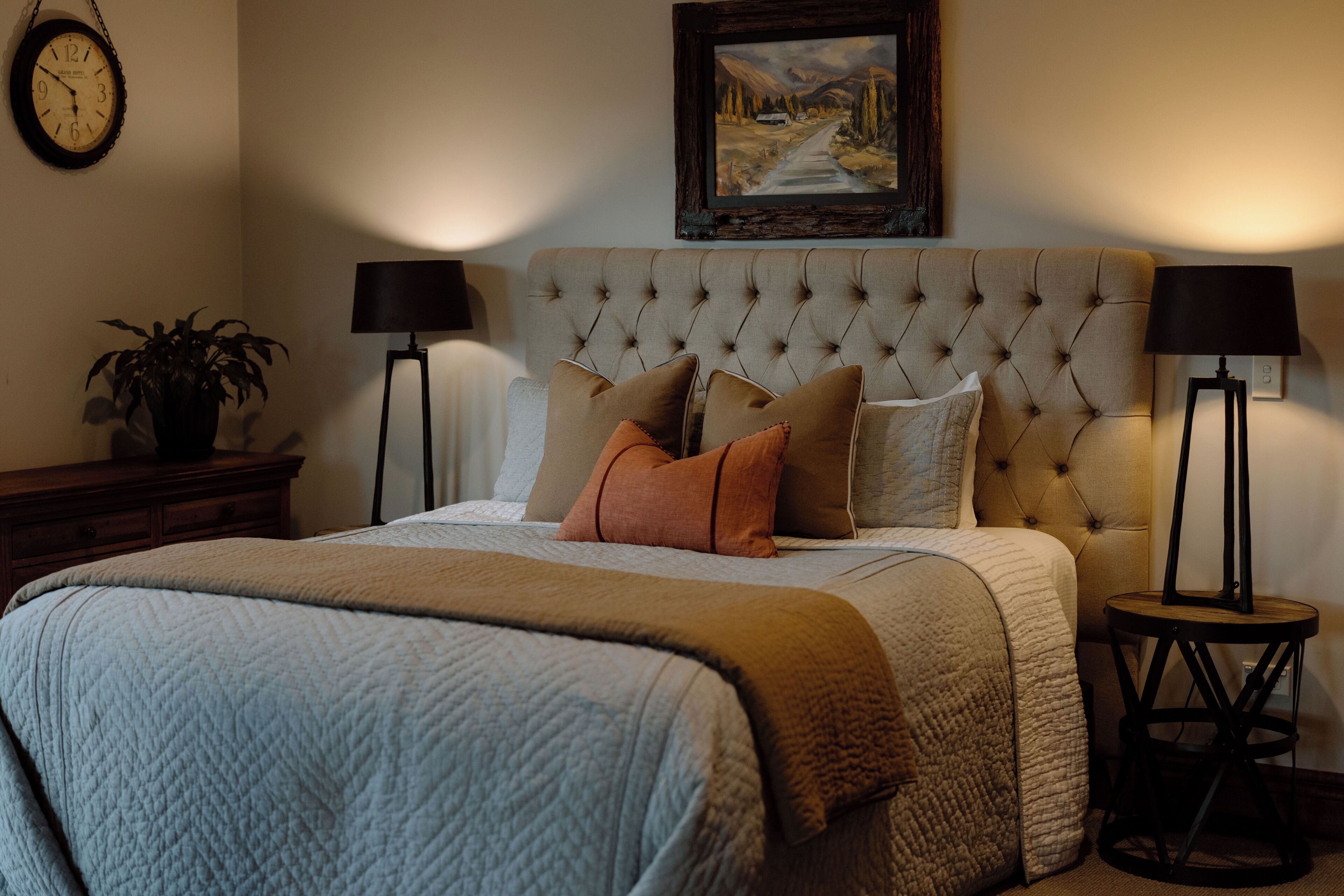 Un dormitorio con cabecera capitonada, una cama grande con muchas almohadas, una mesita de noche con una lámpara y un cuadro en la pared.