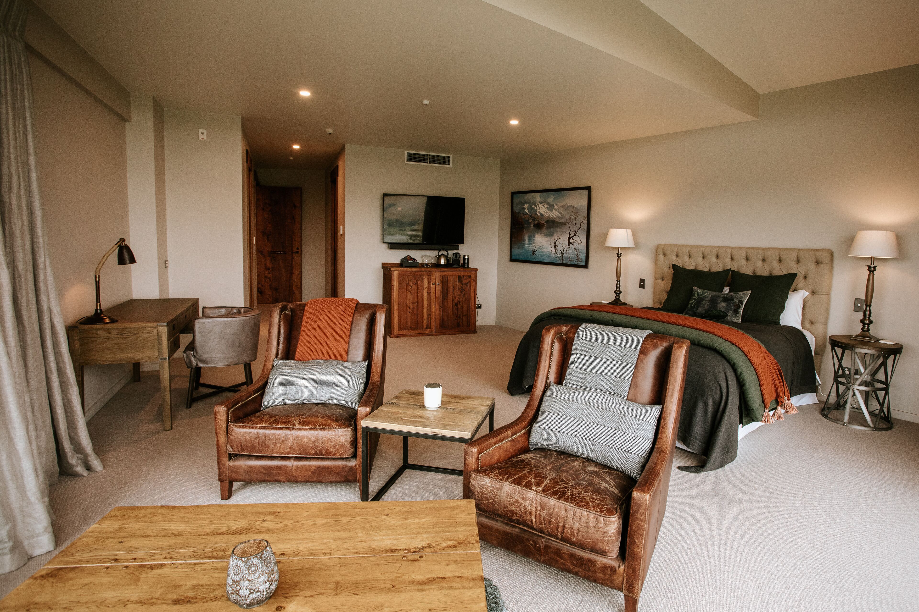 A hotel room with a bed, two leather armchairs, a wooden coffee table, a desk, and a television.