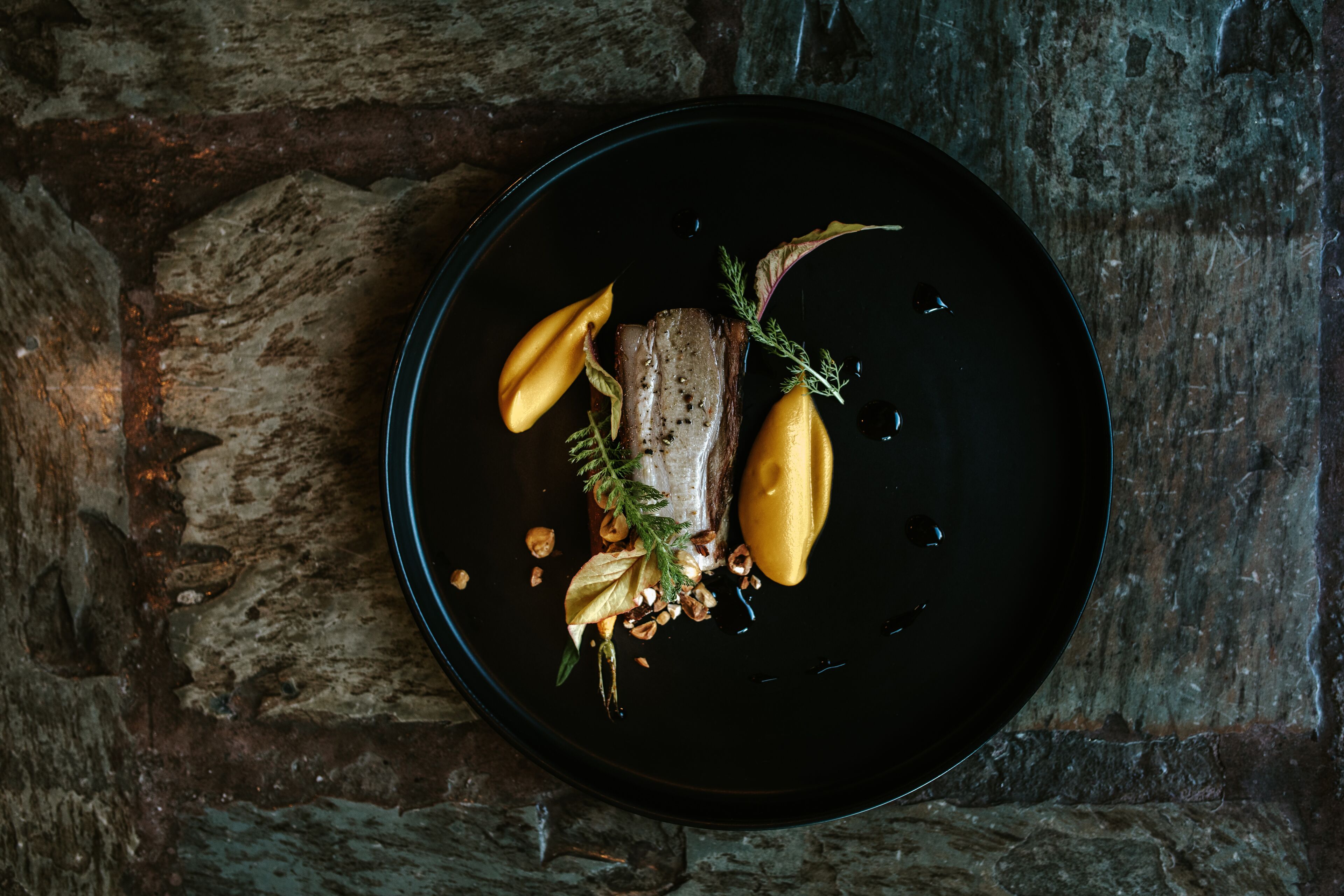 A black plate with a piece of meat, lemon slices, and herbs on a stone surface.