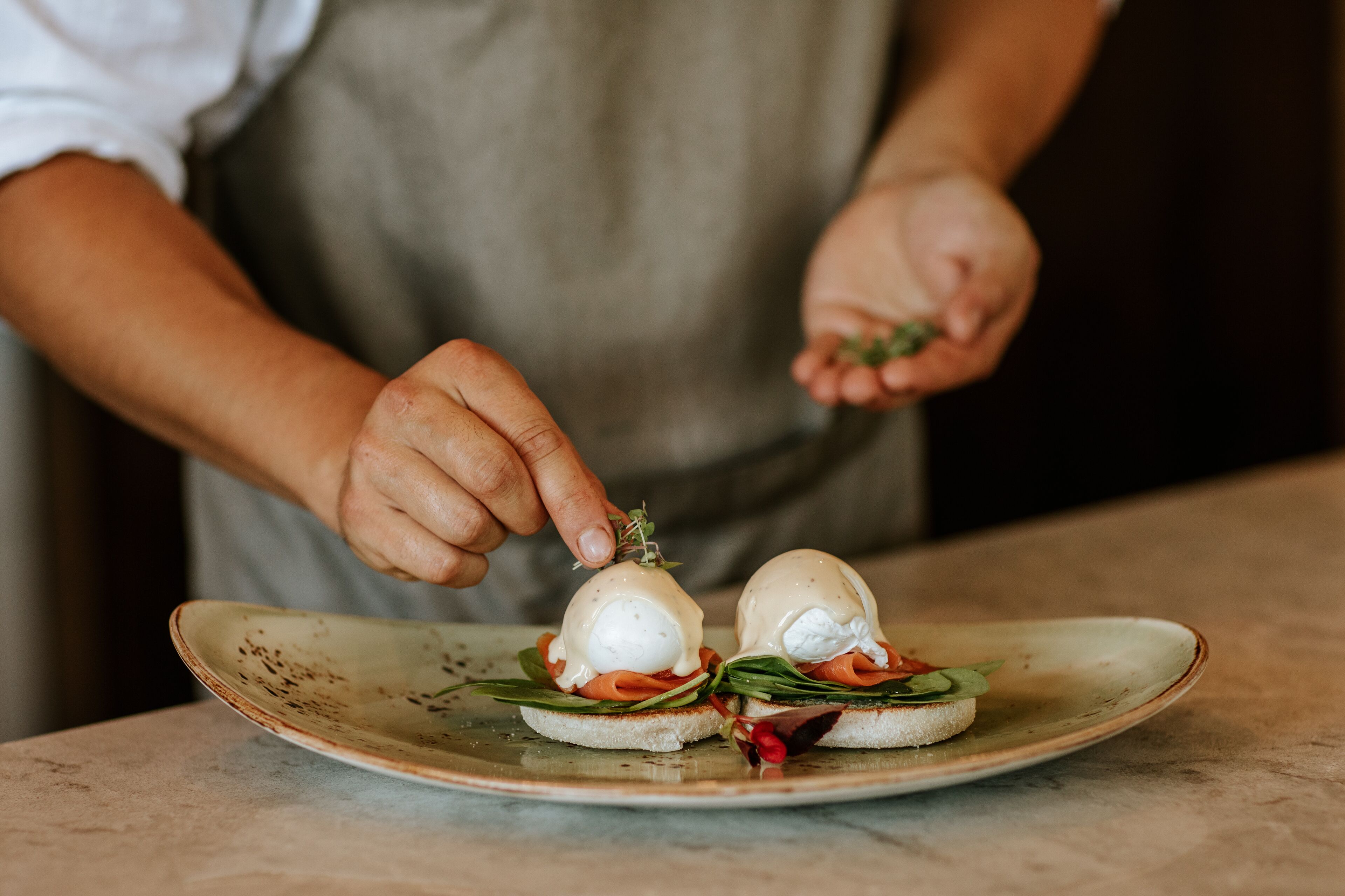A person preparing a dish with poached eggs, smoked salmon, and greens on a ceramic plate.