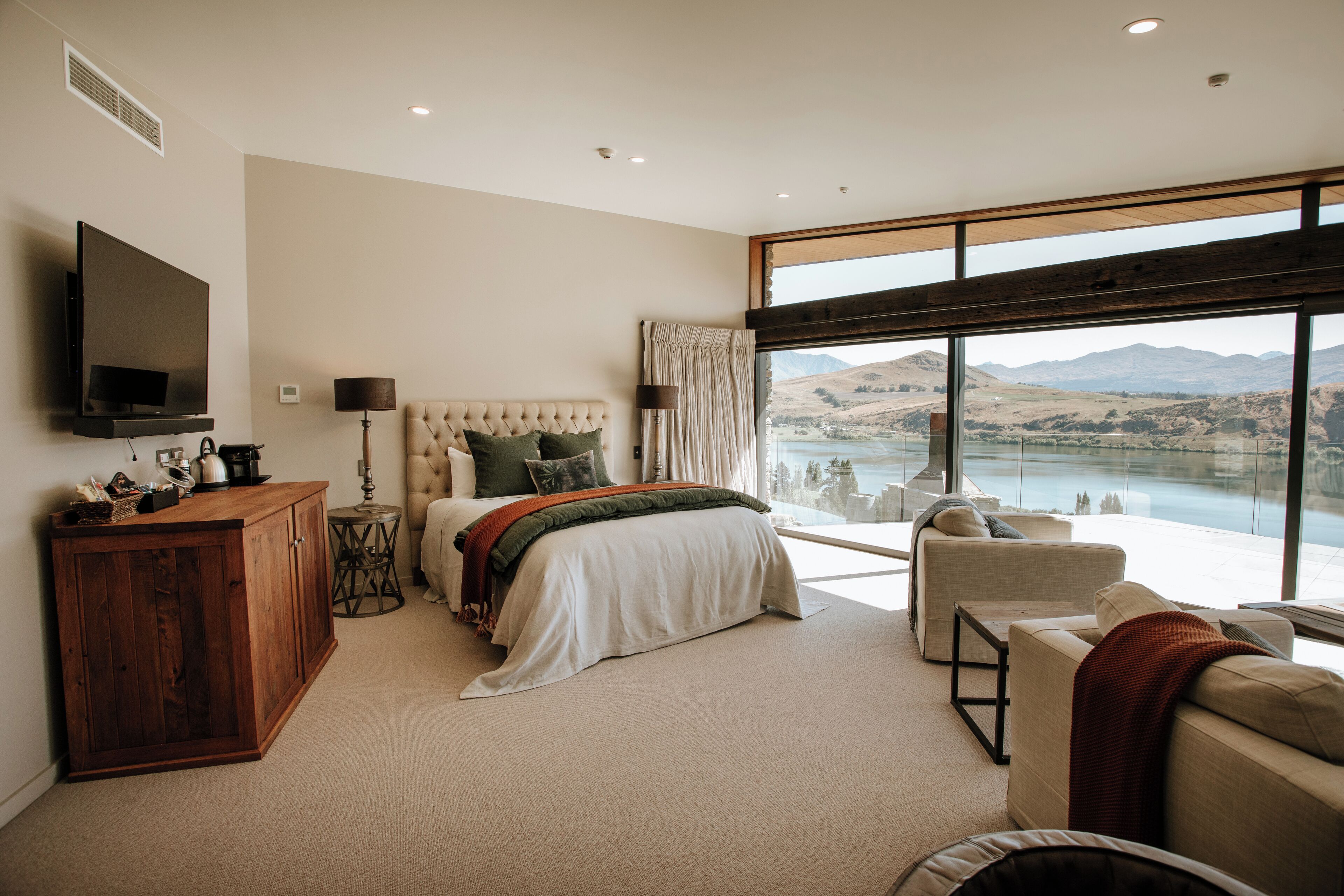 A bedroom with a bed, a TV, a wooden cabinet, a lamp, and a view of a lake and mountains.