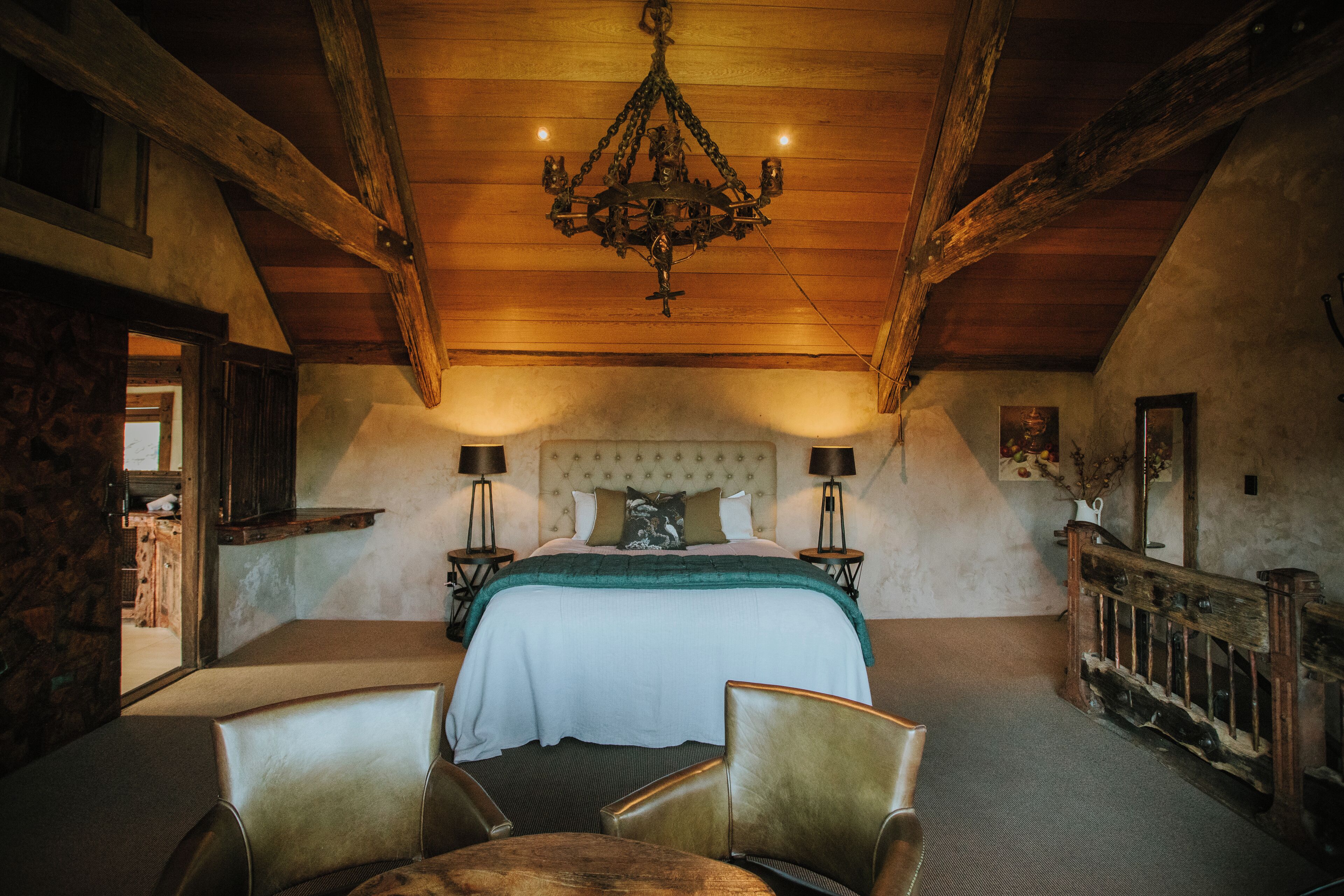 Quarto com cama grande, vigas de madeira, lustre, duas mesas de cabeceira e uma cadeira.