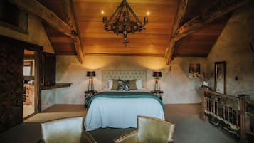Quarto com cama grande, vigas de madeira, lustre, duas mesas de cabeceira e uma cadeira.