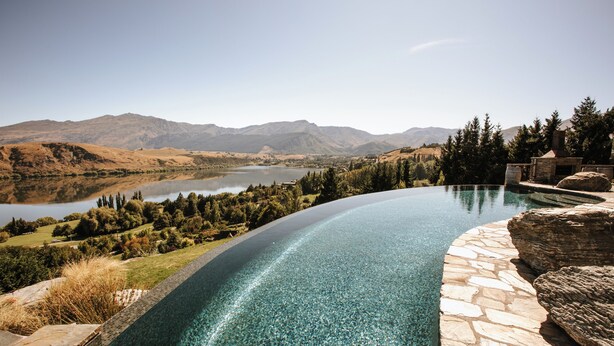Una piscina infinita curvada con vistas despejadas a un lago y montañas al fondo.