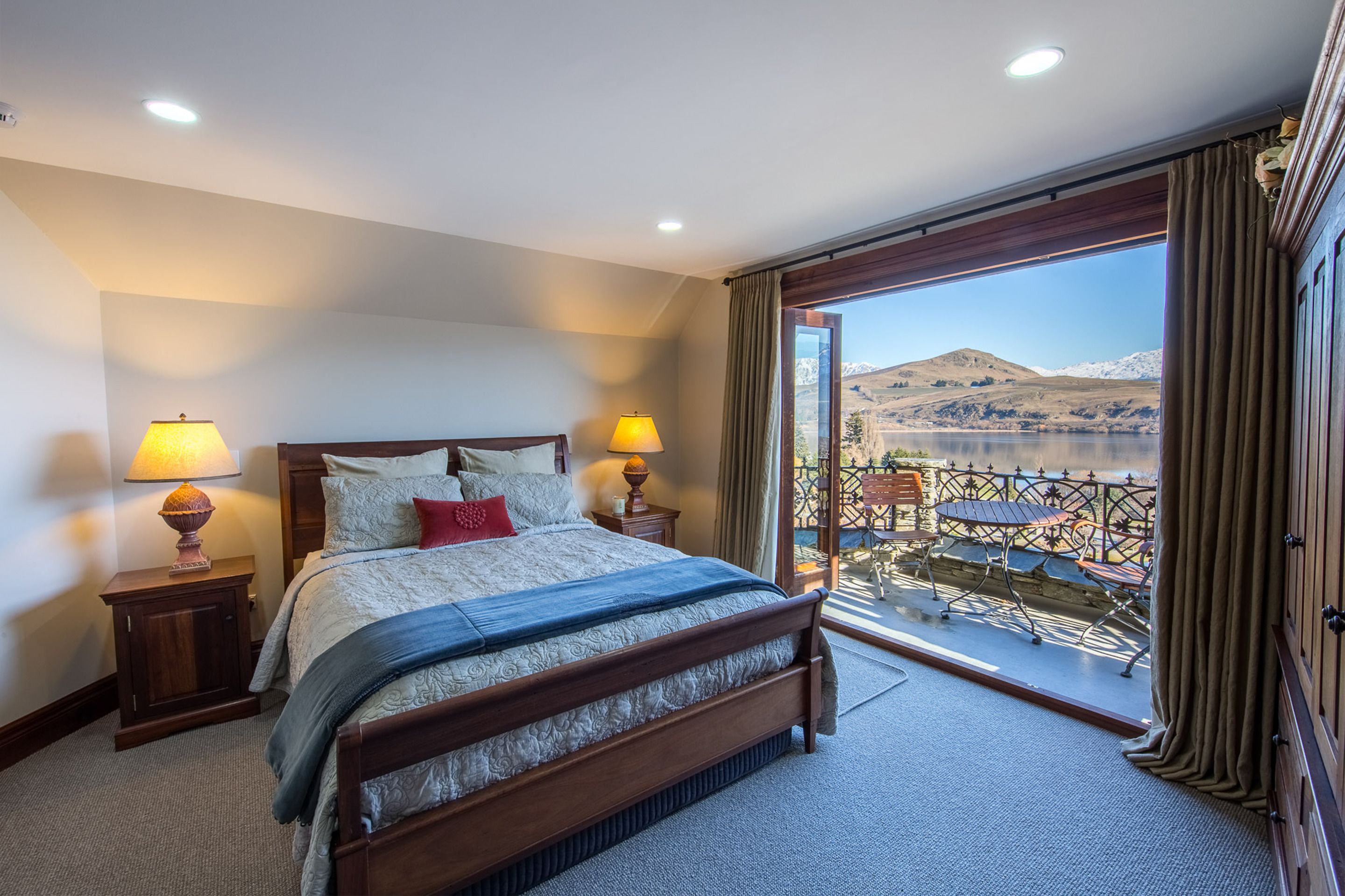 A hotel room with a large bed, two bedside lamps, a view of mountains and a lake through a balcony.