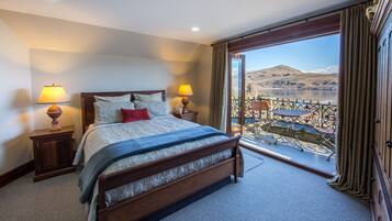 A hotel room with a large bed, two bedside lamps, a view of mountains and a lake through a balcony.