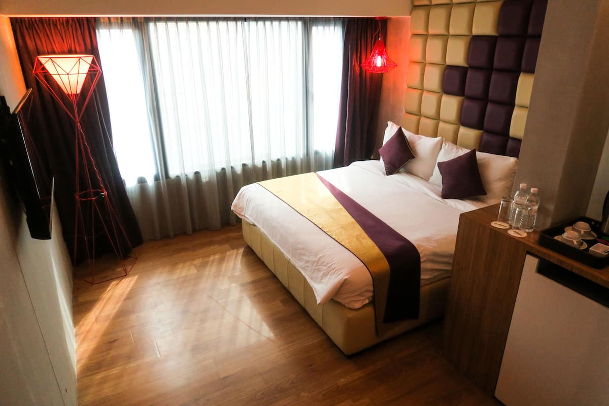 A hotel room with a large bed, a wooden floor, a headboard with a geometric pattern, and a bedside table with a tray of glasses and cups.