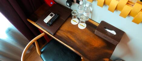 A compact hotel room with a wooden desk, a chair, a small table with a tissue box, two bottles of water, and a phone.
