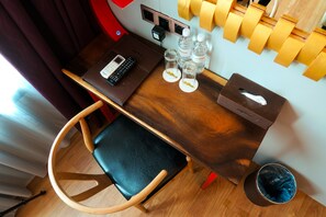 A compact hotel room with a wooden desk, a chair, a small table with a tissue box, two bottles of water, and a phone.