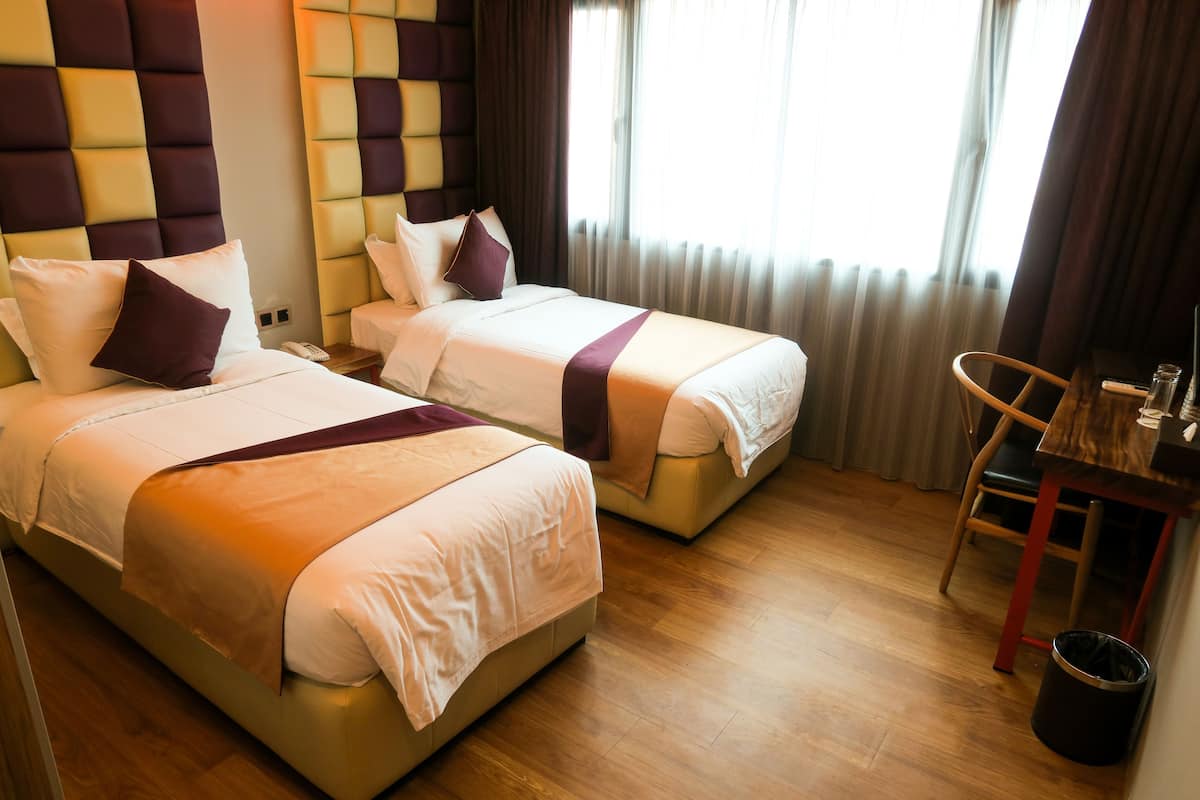 A hotel room with two beds, a wooden floor, a small table, and a trash bin.
