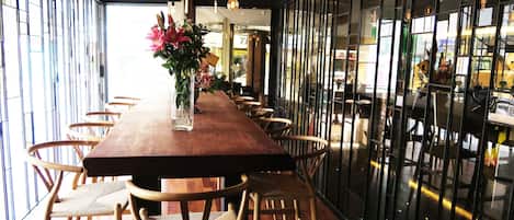 Una larga mesa de madera con sillas de mimbre, un jarrón de flores y lámparas colgantes en un interior moderno con ventanas amplias y paneles de vidrio reflectante.