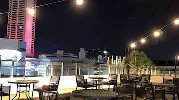 An outdoor deck with tables and chairs, a tall building in the background, and string lights.