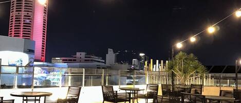 Une terrasse extérieure avec des tables et des chaises, un grand immeuble en arrière-plan et des guirlandes lumineuses.