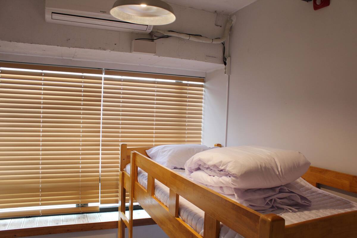A single bed with a wooden headboard, a window with blinds, and a ceiling-mounted air conditioning unit.