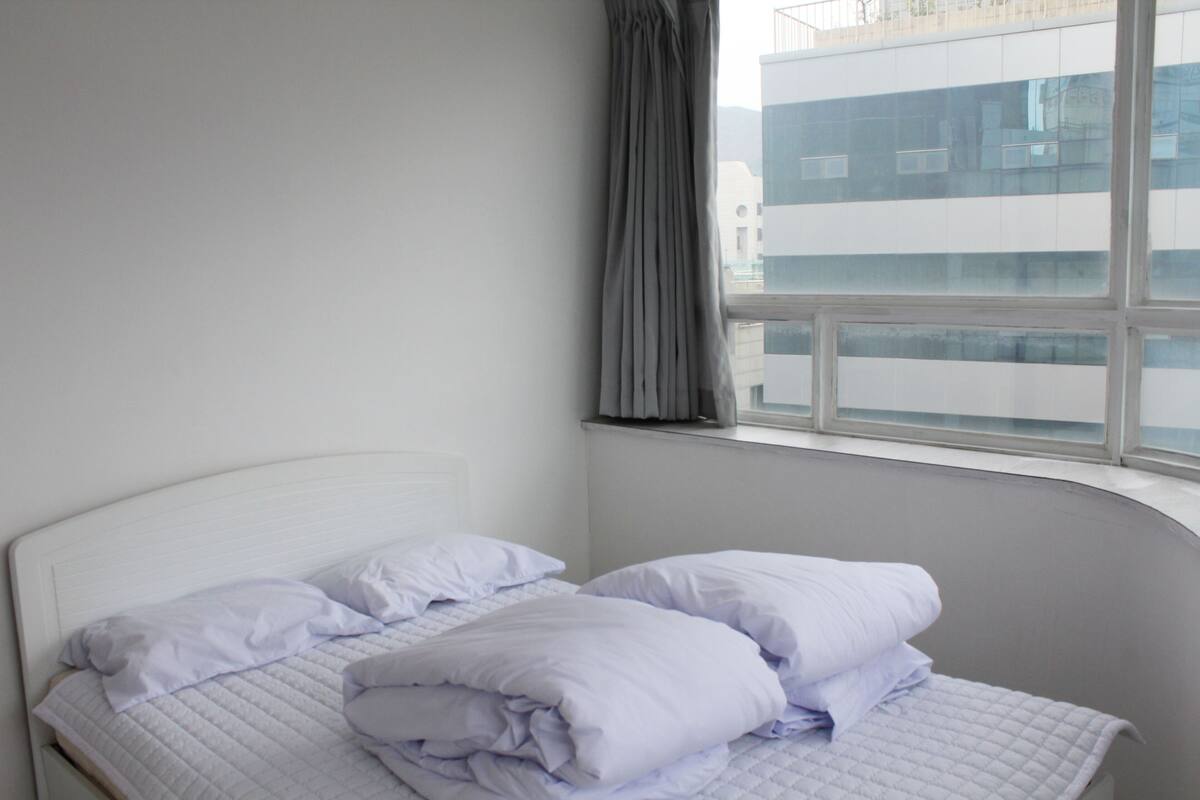 A neatly made bed with white bedding in a room with a large window and a view of a building outside.