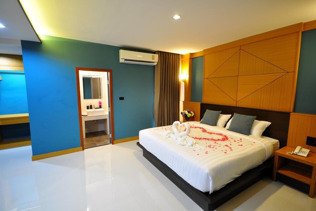 A modern hotel room with a large bed, a wooden headboard, a bathroom with a mirror, and an air conditioning unit.