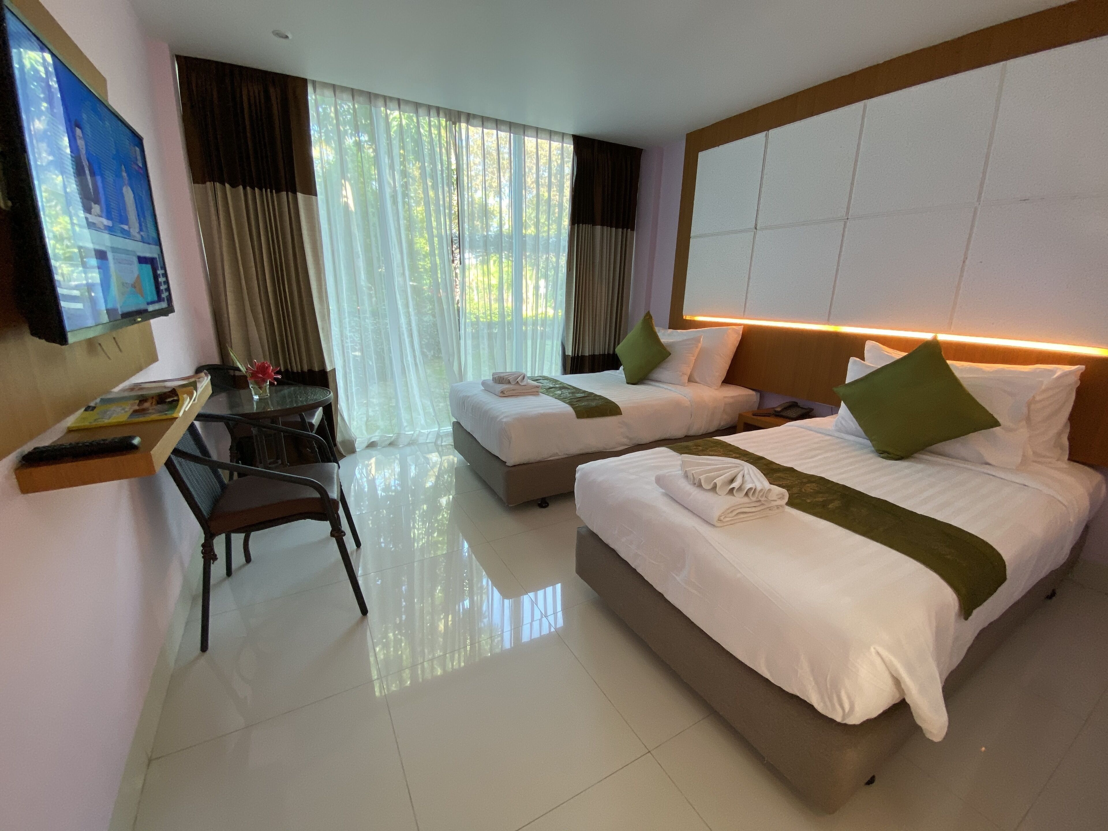 Ein Hotelzimmer mit zwei Betten, einem Schreibtisch mit Stuhl, einem Fernseher und einem großen Fenster mit Vorhängen.