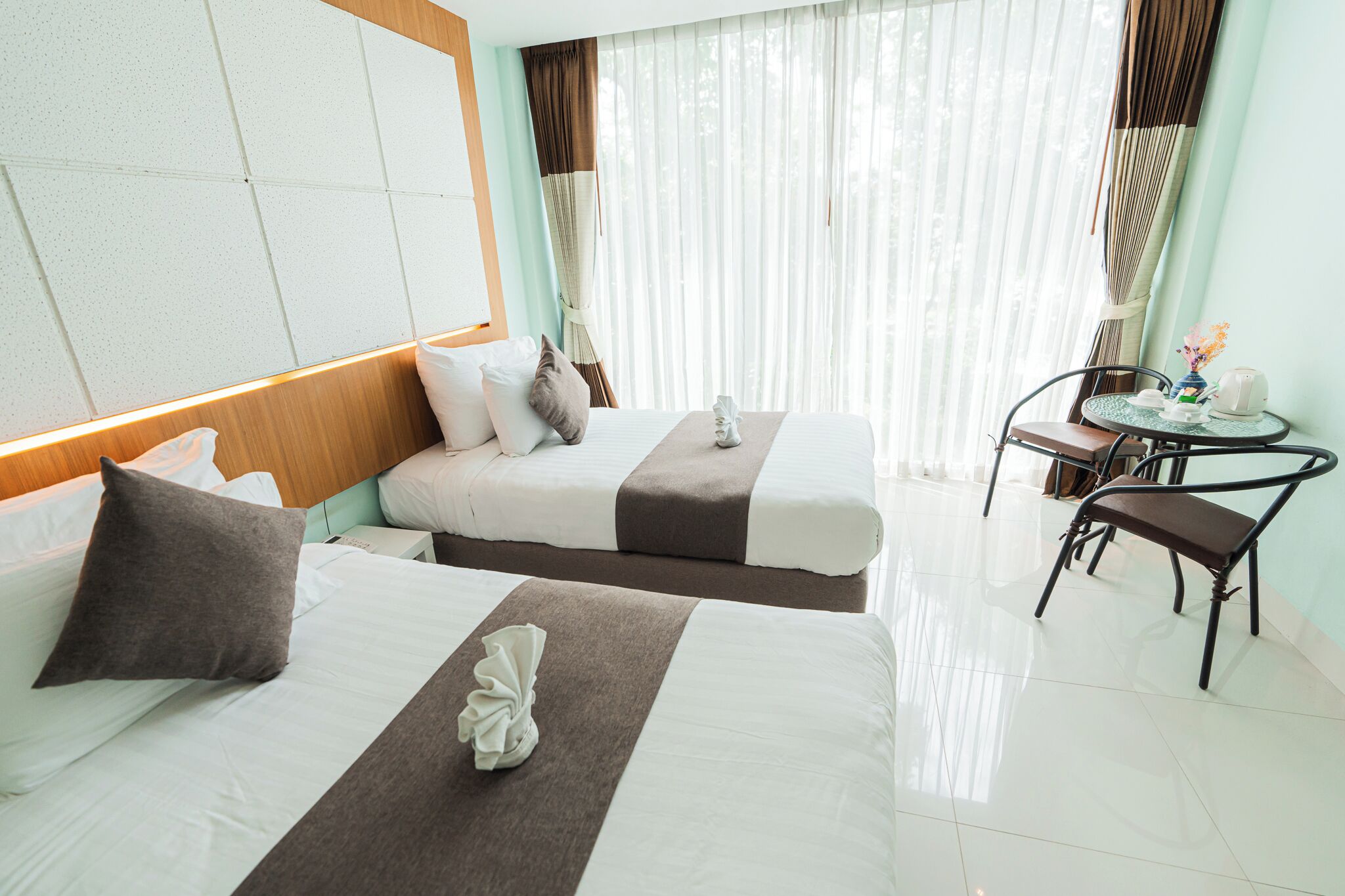 A hotel room with two beds, a small table with a chair, and a window with curtains.