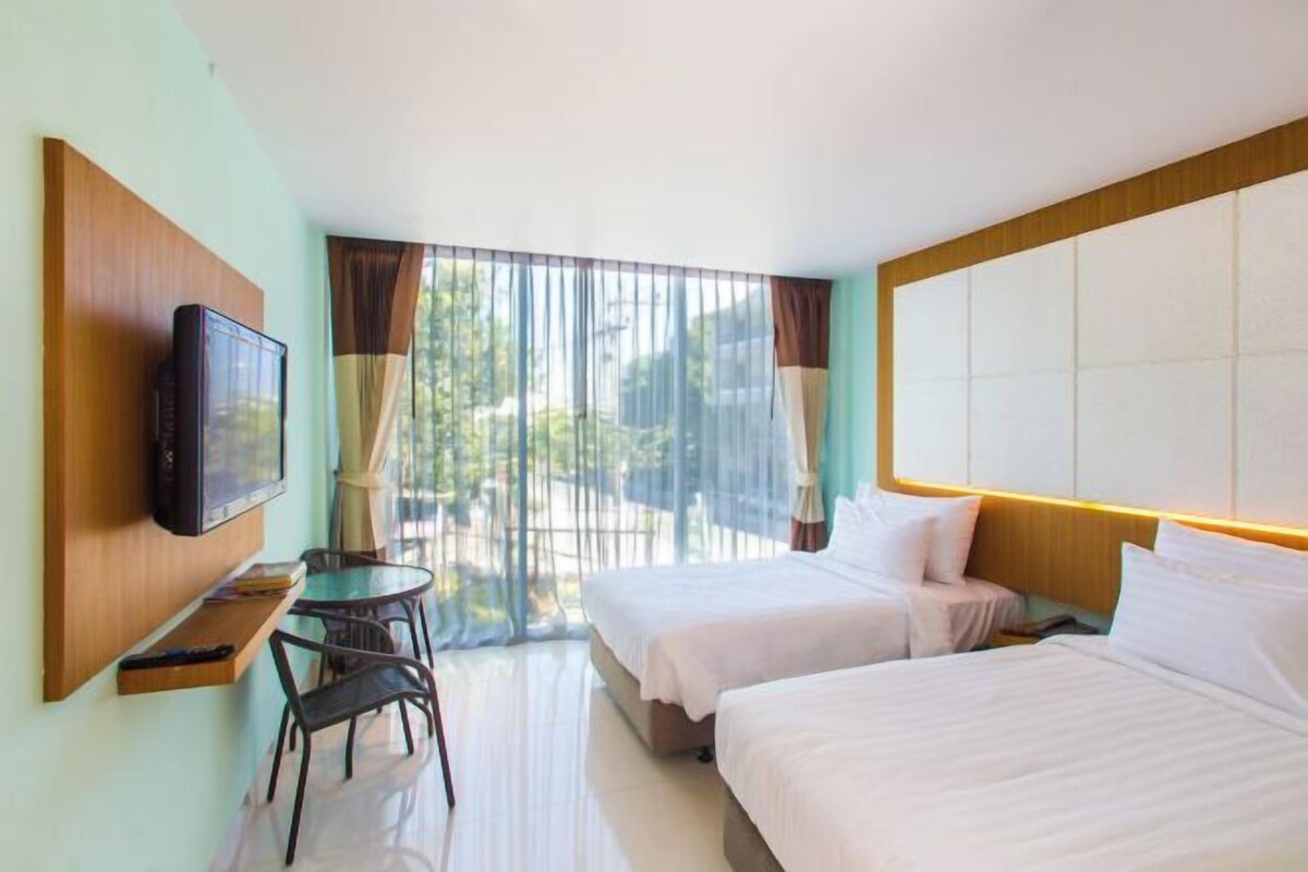 A hotel room with two beds, a TV mounted on the wall, a small table with chairs, and a large window with curtains.