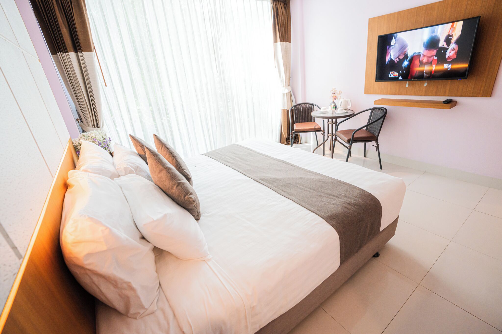 A hotel room with a large bed, a TV mounted on the wall, a small table with chairs, and a window with curtains.