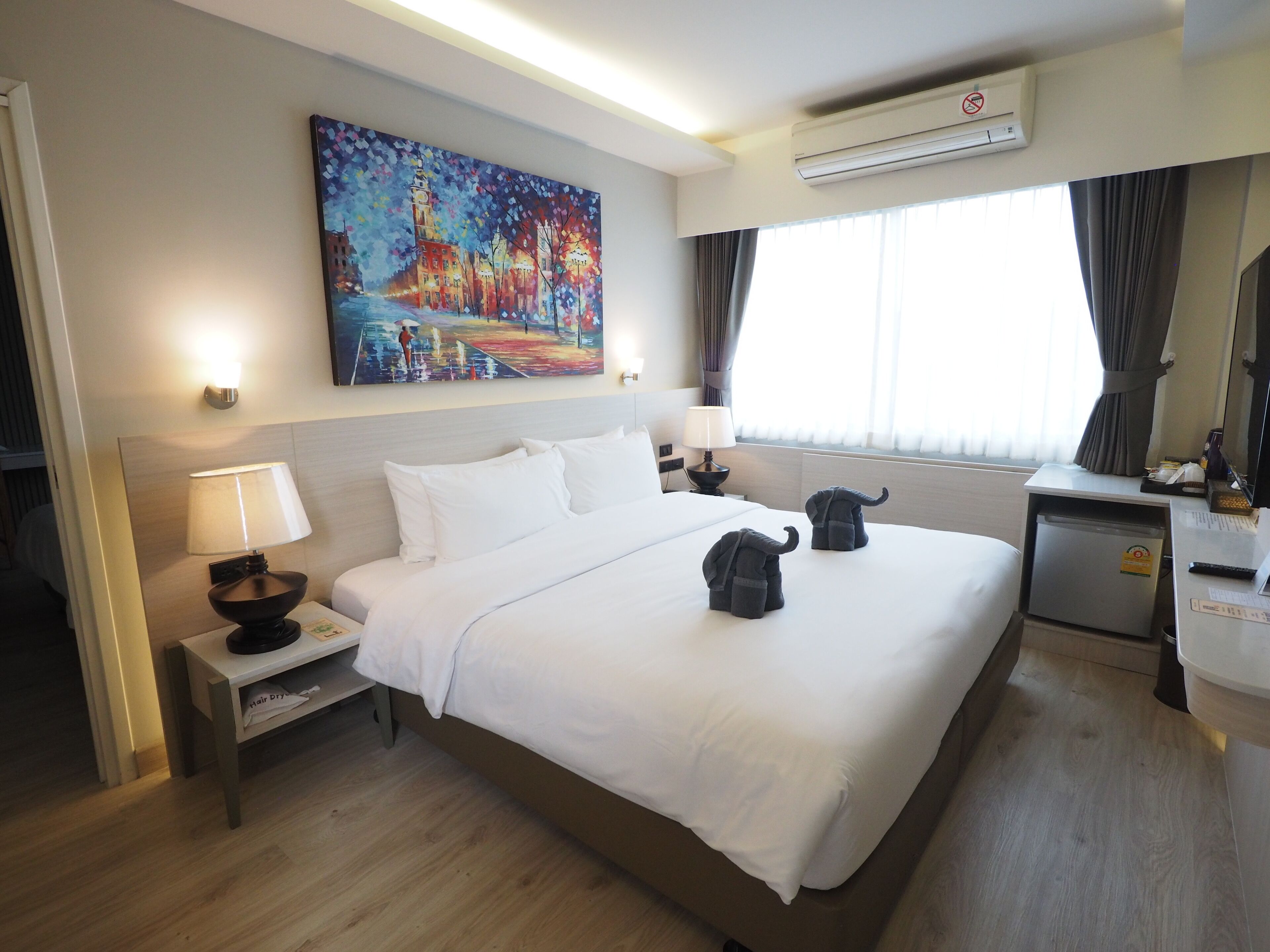 Habitación de hotel con una cama grande, dos mesitas de noche con lámparas, un escritorio con silla, un refrigerador pequeño y un cuadro en la pared.