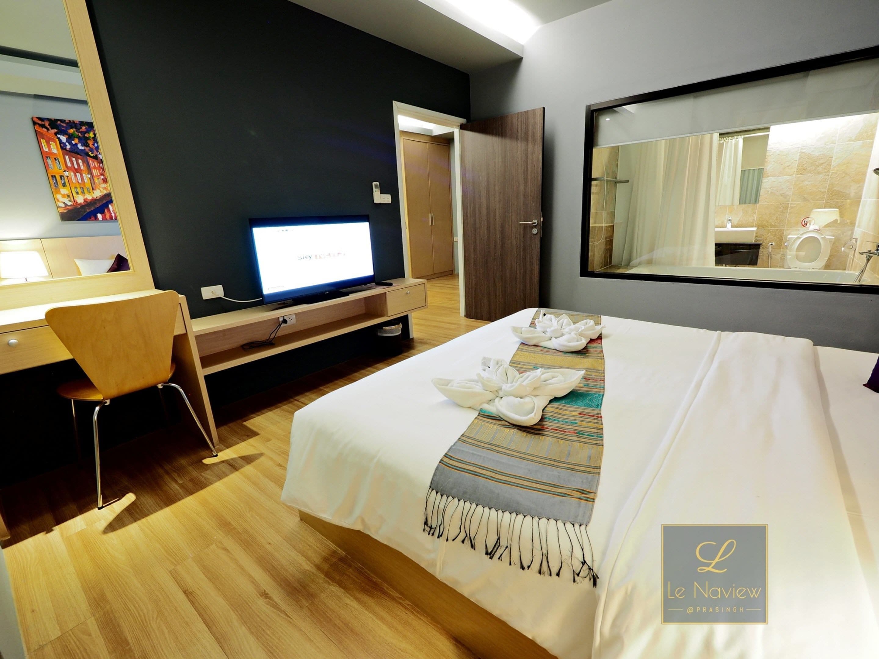 Habitación de hotel con una cama, un escritorio, una silla, un televisor y un espejo grande.
