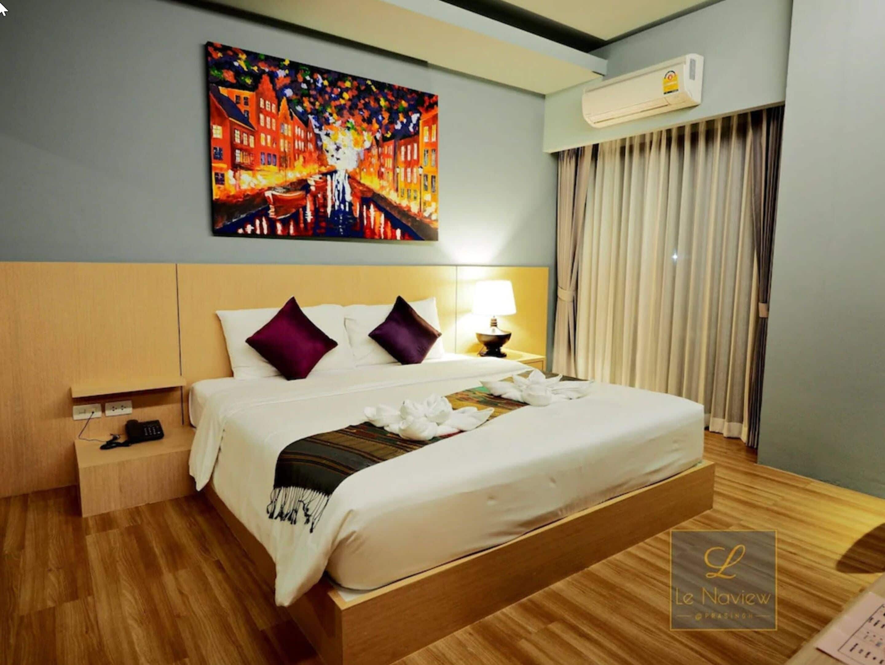 A hotel room with a large bed, a painting on the wall, an air conditioning unit, and a window with curtains.