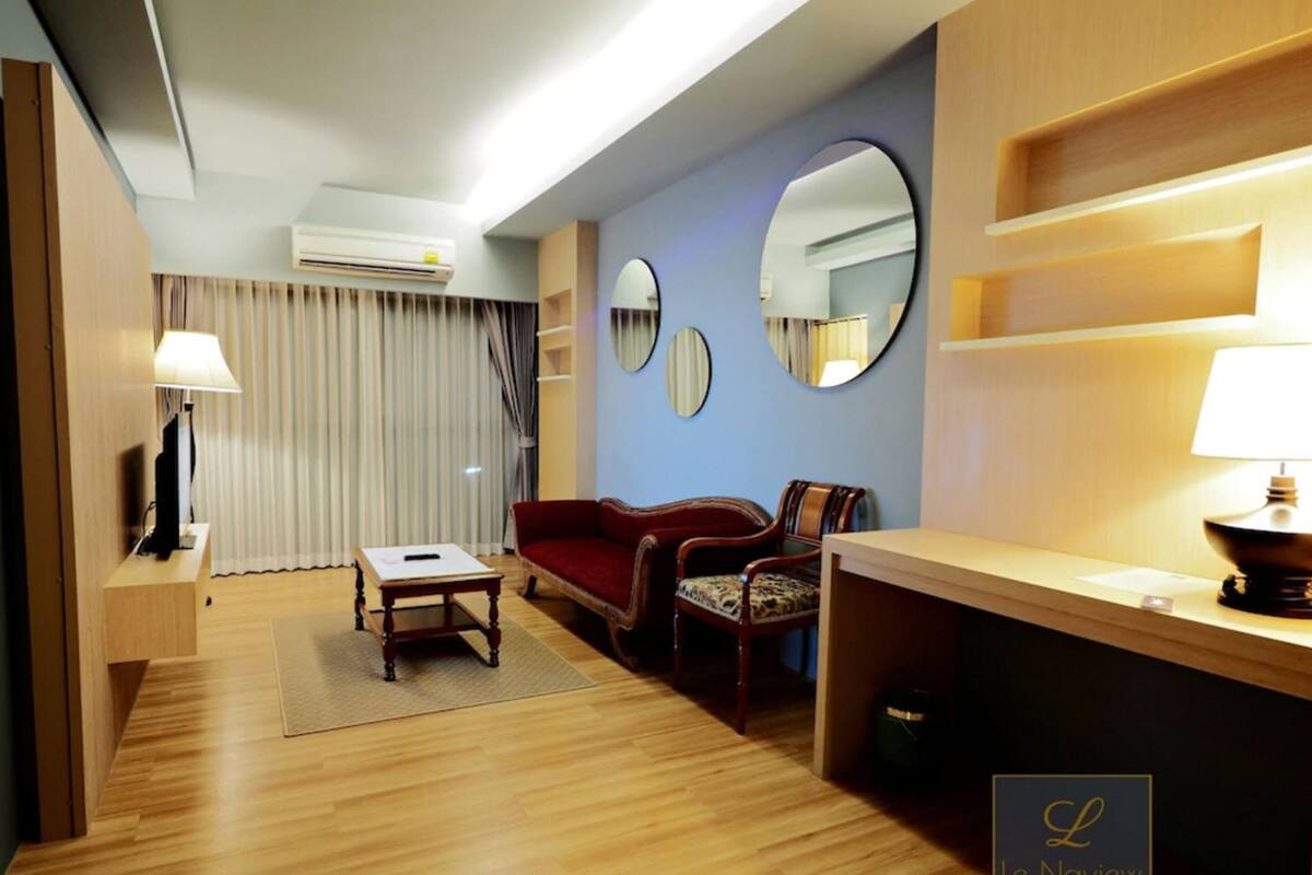 Habitación de hotel con suelo de madera, un sofá rojo, una mesita, un televisor y un escritorio con lámpara. Hay dos espejos en la pared y un equipo de aire acondicionado sobre el sofá.