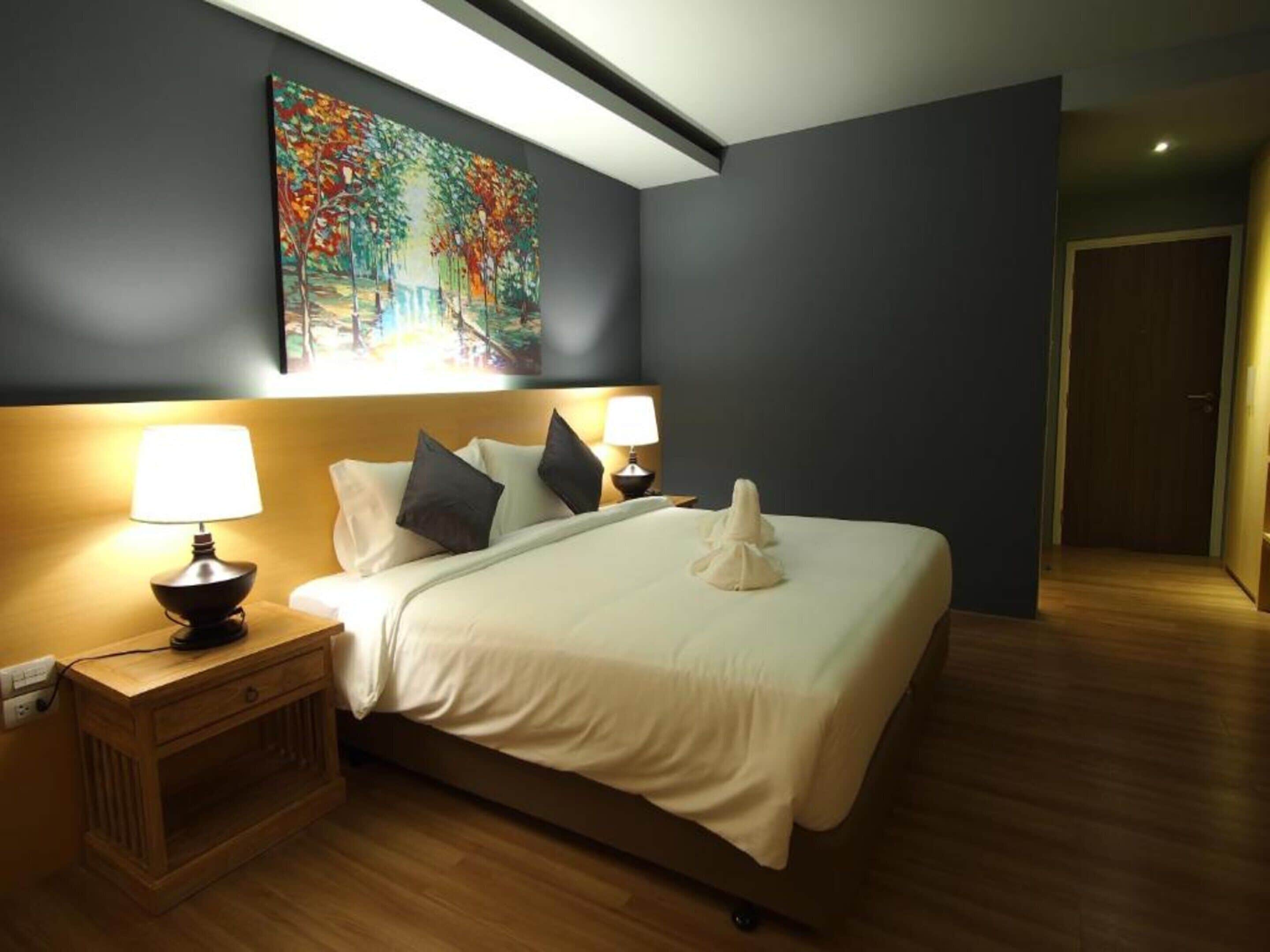 A hotel room with a large bed, two bedside lamps, a wooden nightstand, a painting on the wall, and a door in the background.