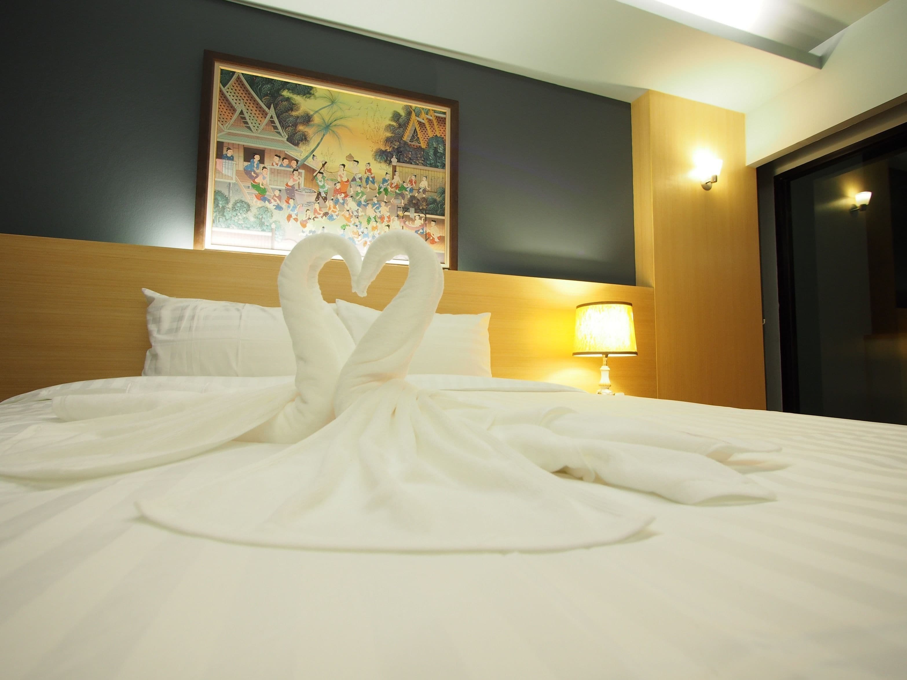 A hotel room with a bed featuring a heart-shaped towel arrangement, a painting on the wall, and a nightstand with a lamp.