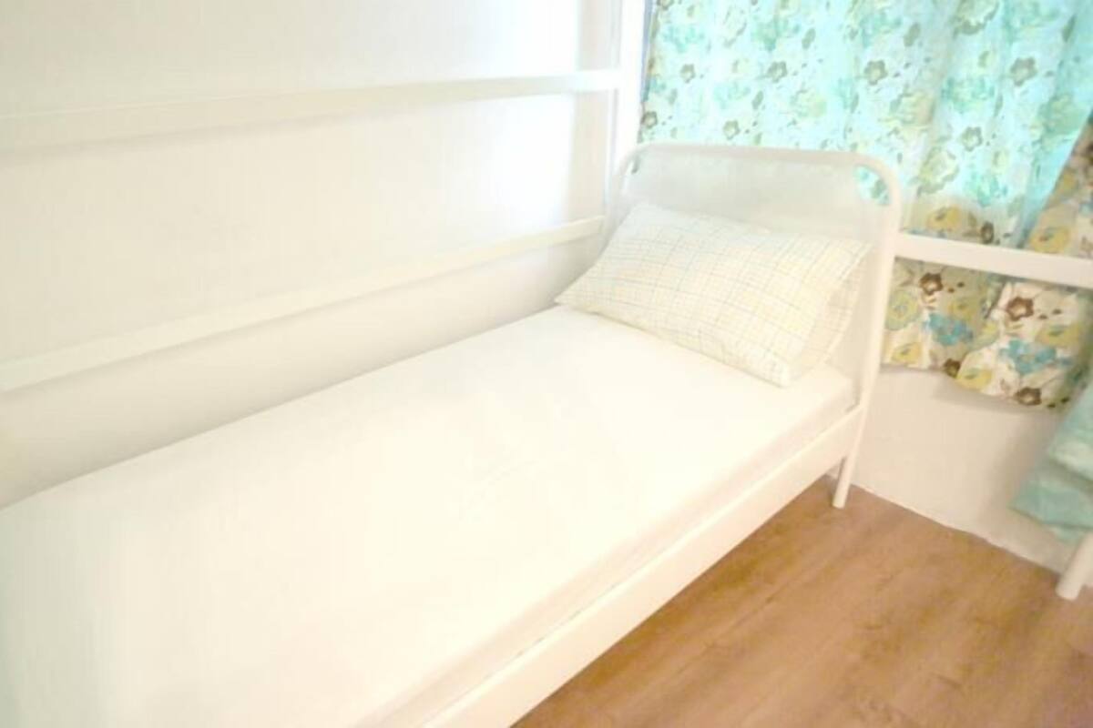 A small, single-bed room with a white mattress and headboard, light wooden flooring, and floral-patterned curtains.