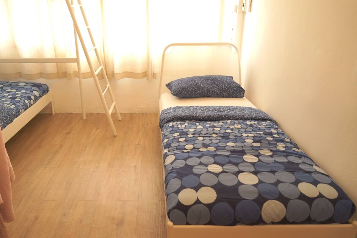 A small, single-bed room with a wooden floor, a ladder, and a window with curtains.