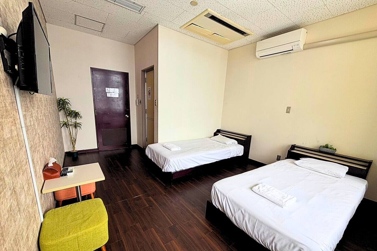Habitación de hotel con dos camas individuales, un escritorio con silla, televisor montado en la pared y equipo de aire acondicionado.