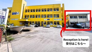 A yellow building with multiple floors, signs indicating a reception area, and several parked cars in front. - My Place - Hostel (Naha)