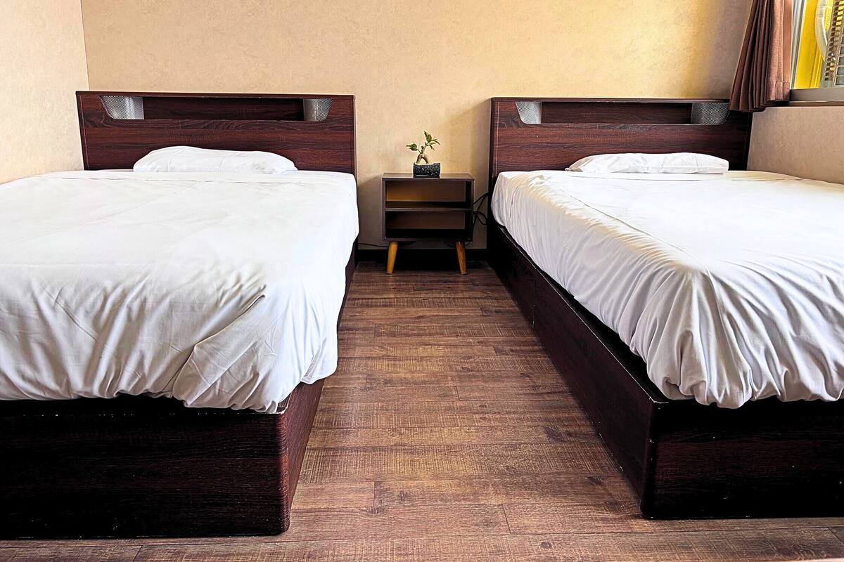 Una habitación de hotel con dos camas individuales, una mesita de noche pequeña con lámpara y una ventana con cortinas.
