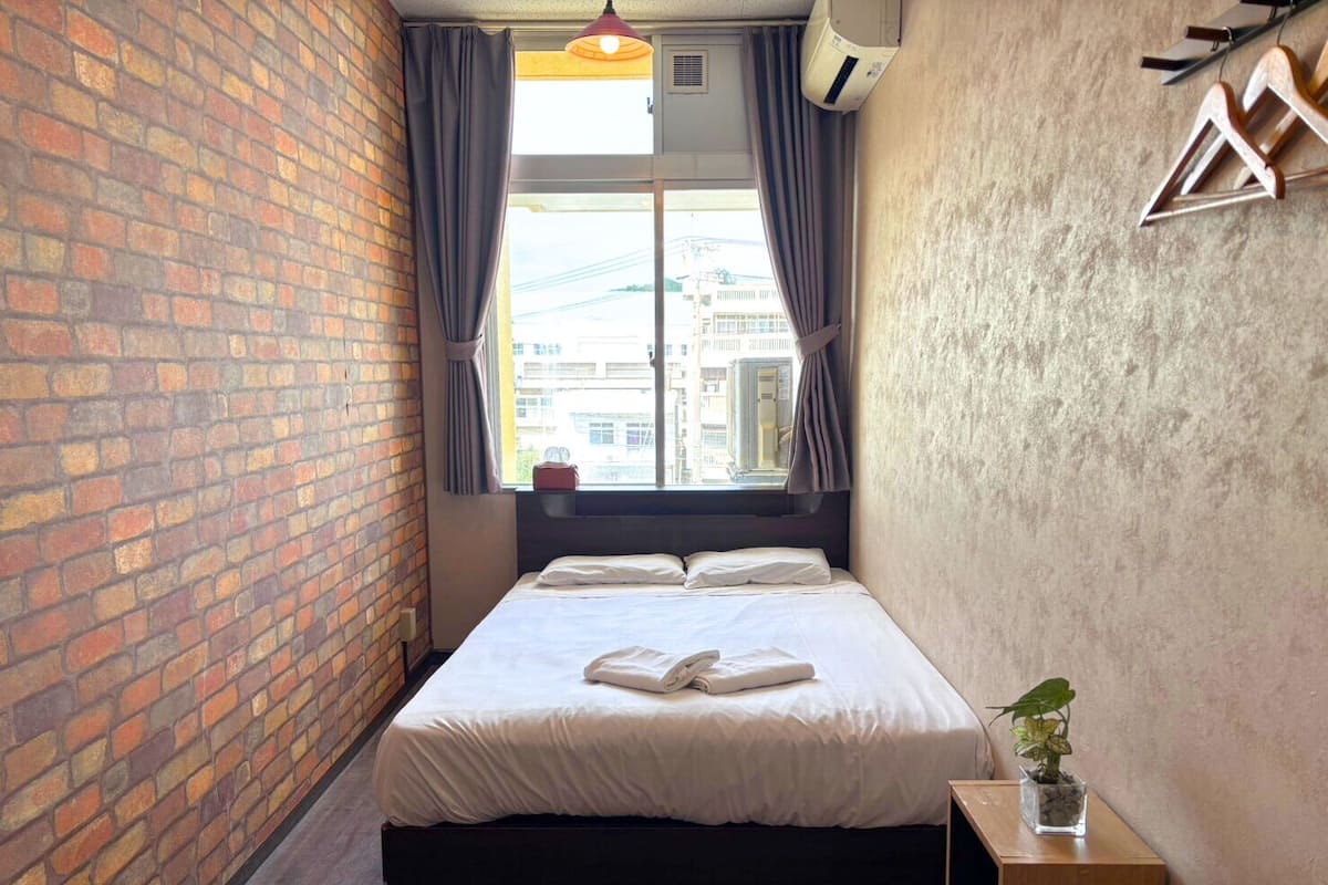 Una habitación de hotel pequeña y moderna con pared de ladrillo, un ventanal con cortinas, una cama con sábanas blancas y una planta pequeña sobre una mesita de noche.