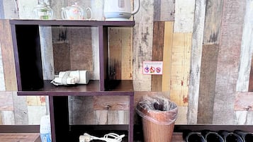 Una estantería de madera con un hervidor, un frasco de vidrio y una botella de plástico. Hay un cubo de basura y un par de zapatillas en el suelo.