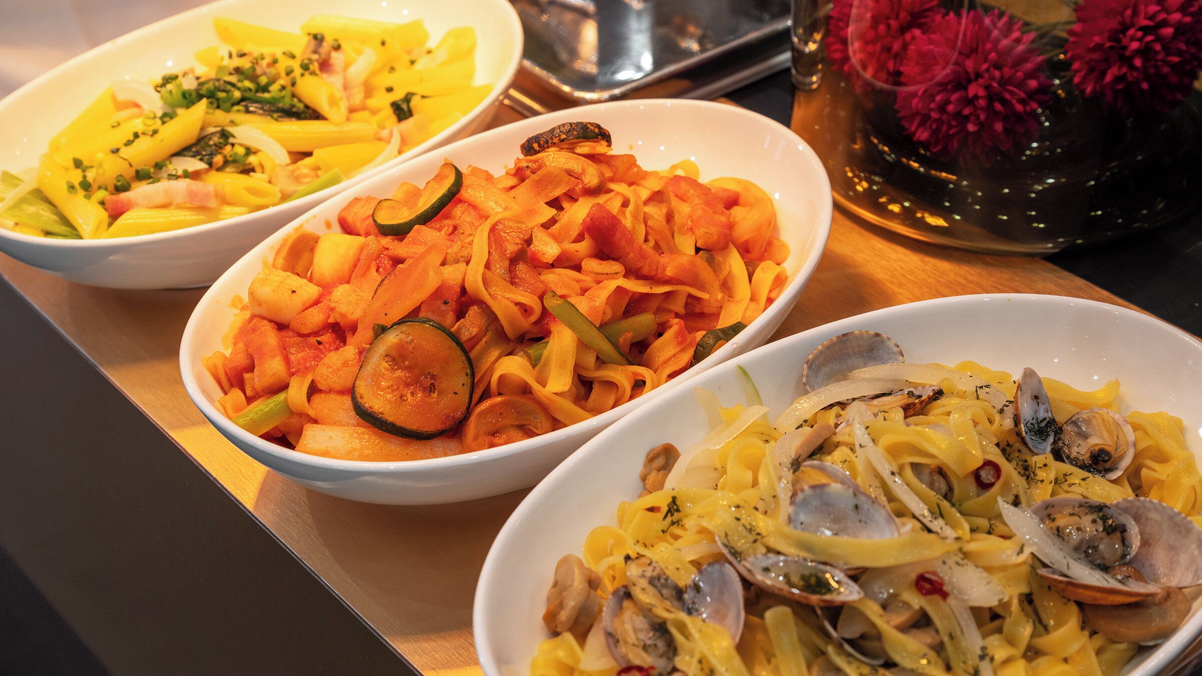 Tres fuentes de platos de pasta con verduras y mariscos sobre una mesa.
