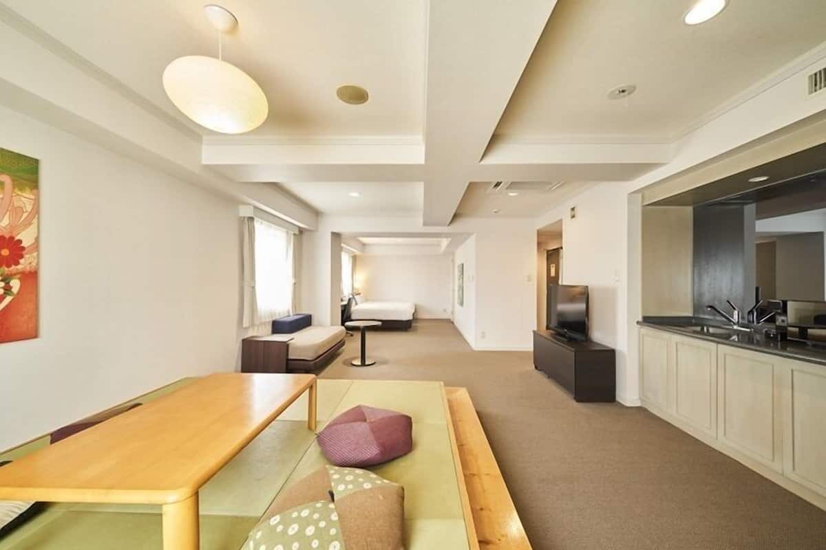 A modern hotel room with a wooden dining table, a sofa, a TV, and a kitchenette.