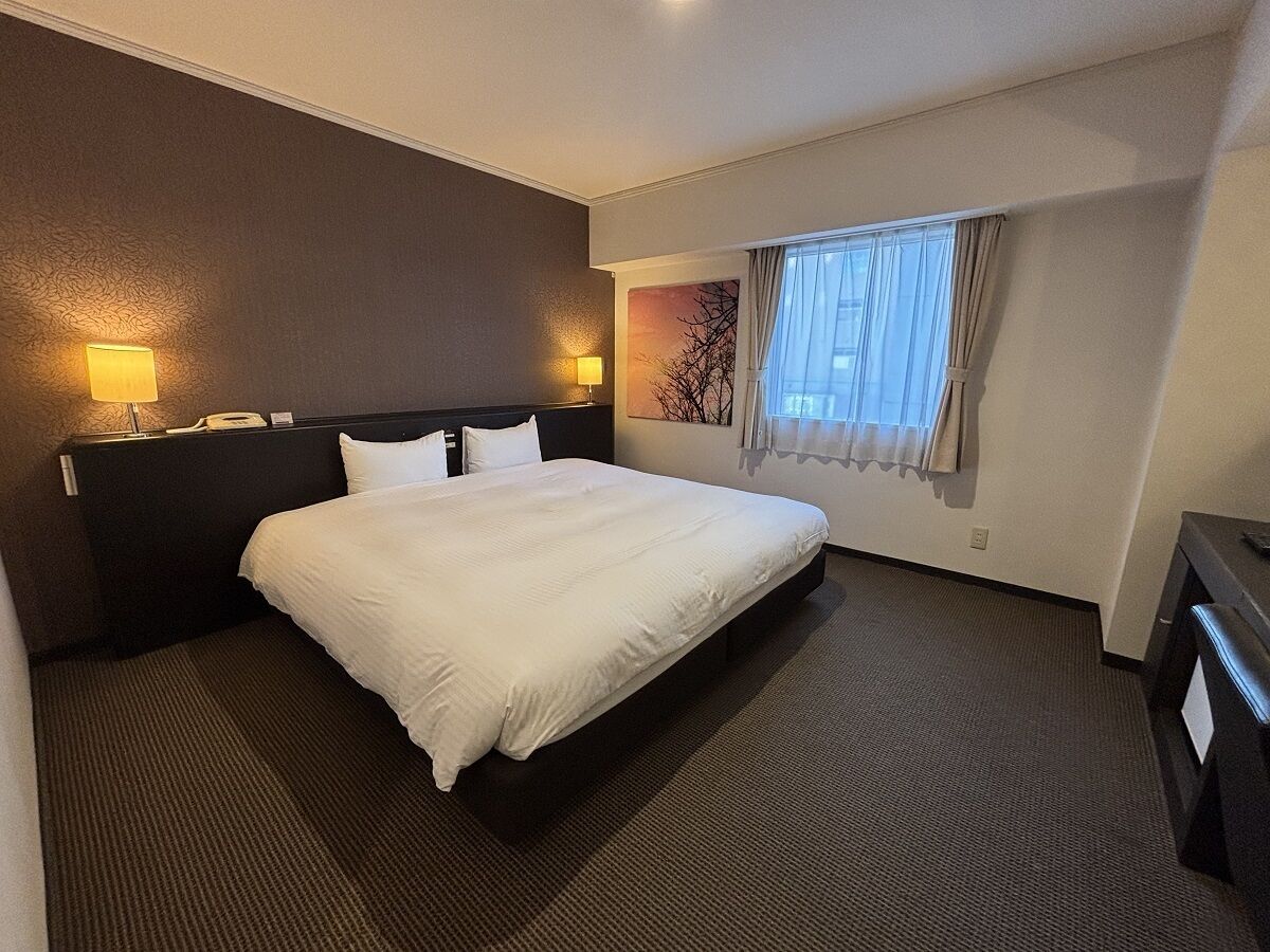 A hotel room with a large bed, two bedside lamps, a window with curtains, and a wall-mounted artwork.