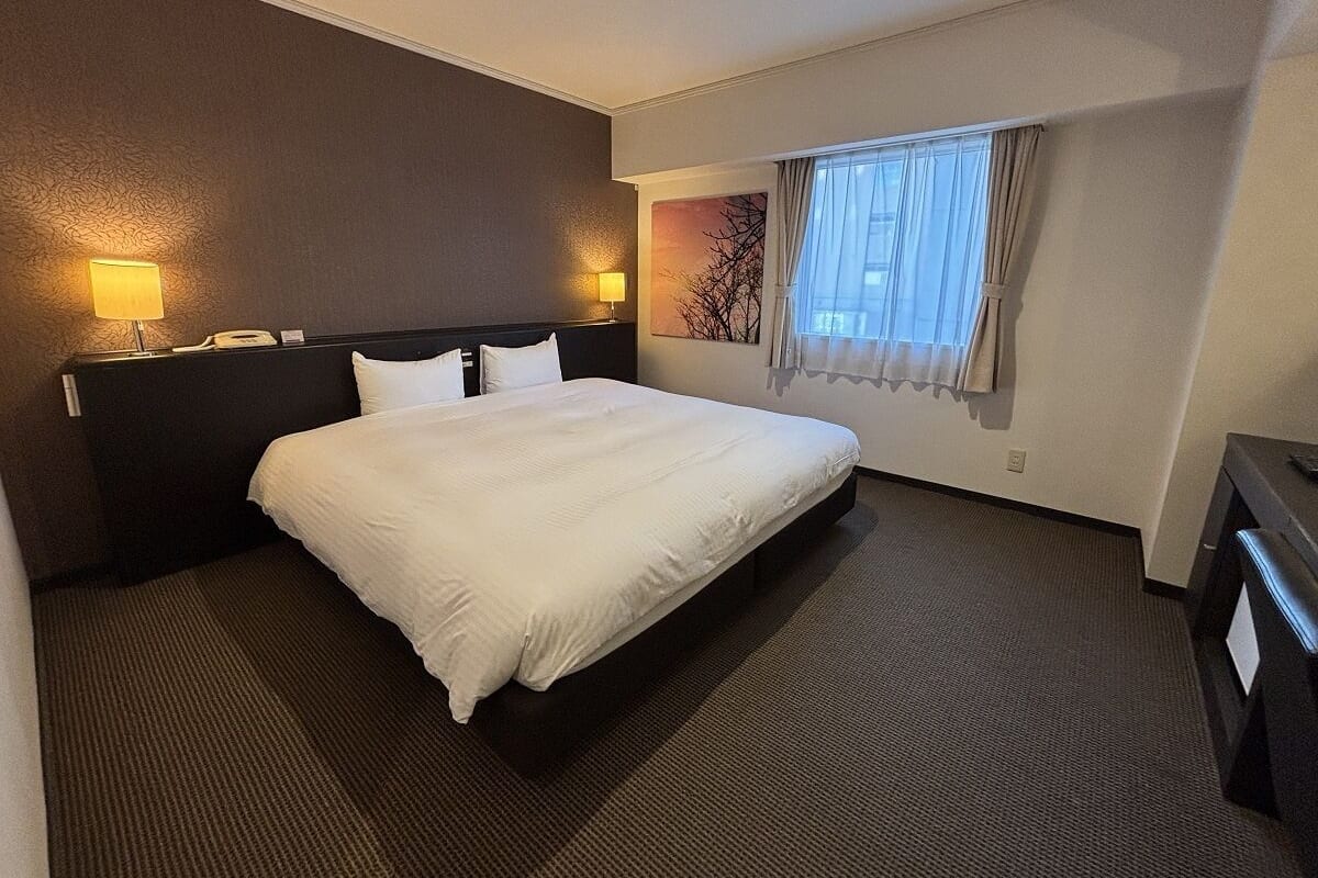 A hotel room with a large bed, two bedside lamps, a window with curtains, and a wall-mounted artwork.