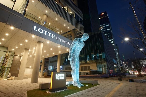 Exterior del hotel Lotte City Hotels, con una llamativa escultura iluminada de una figura humana.