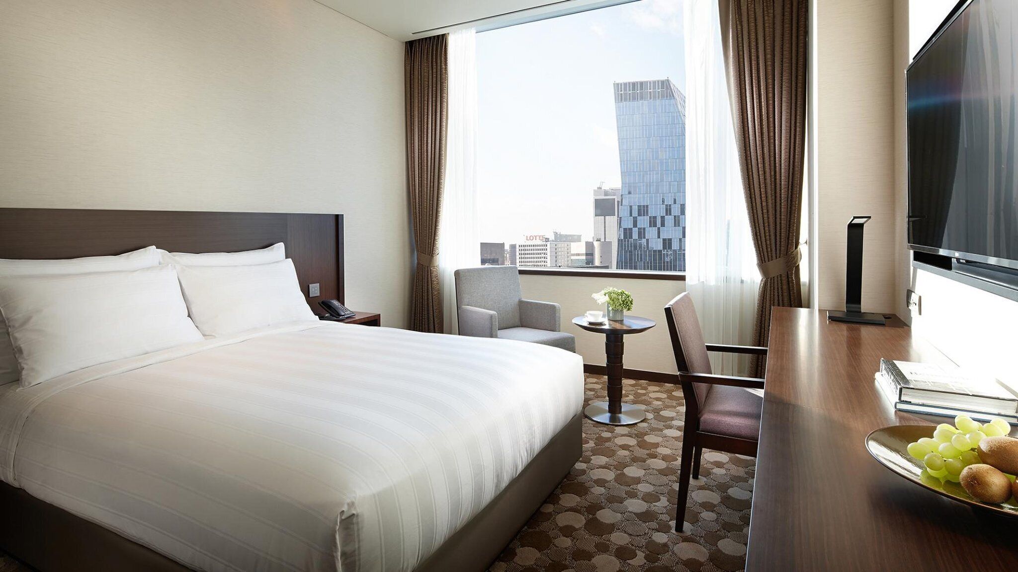 A hotel room with a large bed, a desk with a chair, a small table with a plant, and a view of the cityscape.
