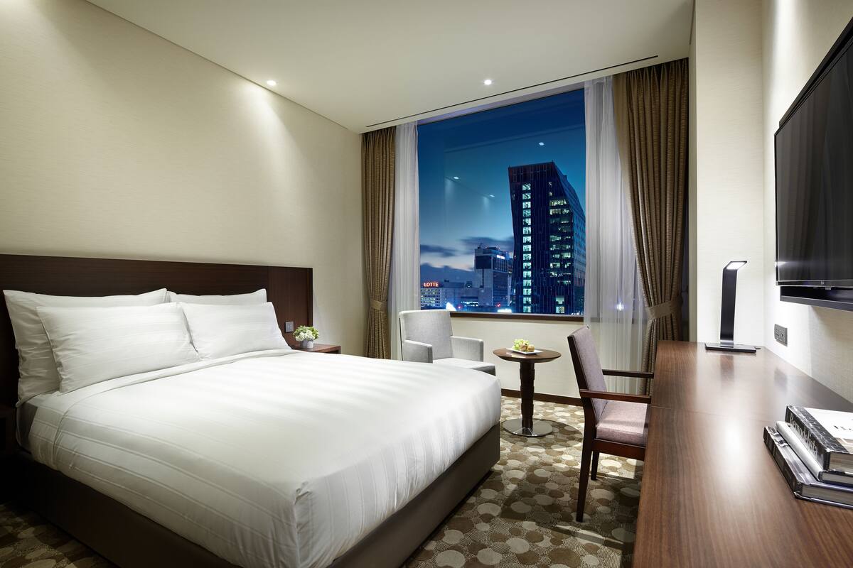 A hotel room with a large bed, a desk with a chair, a small table with a plant, and a TV on the wall.