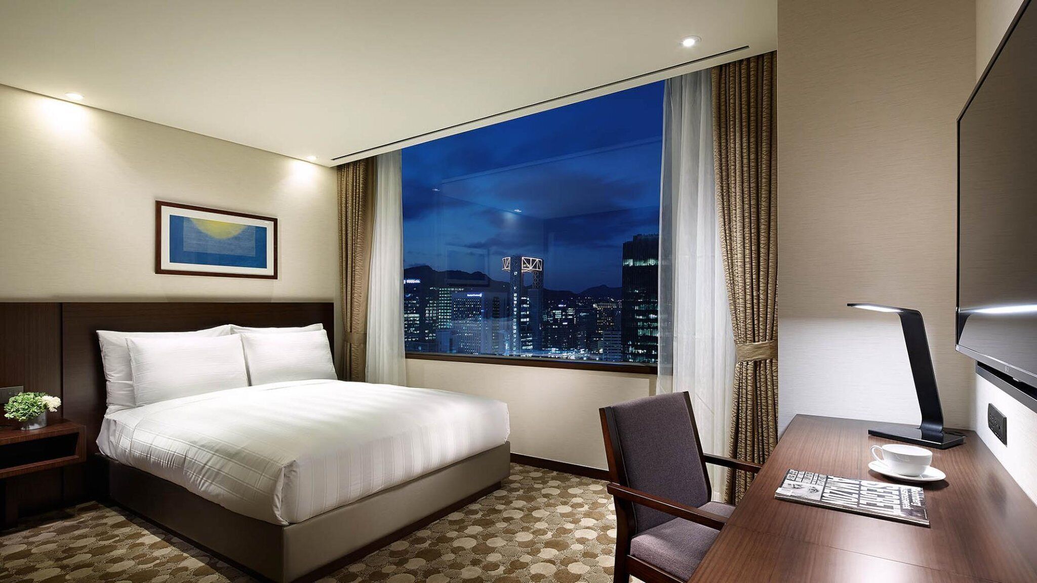 A hotel room with a bed, a desk, a chair, a TV, and a window with a city view.