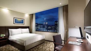 A hotel room with a bed, a desk, a chair, a TV, and a window with a city view.